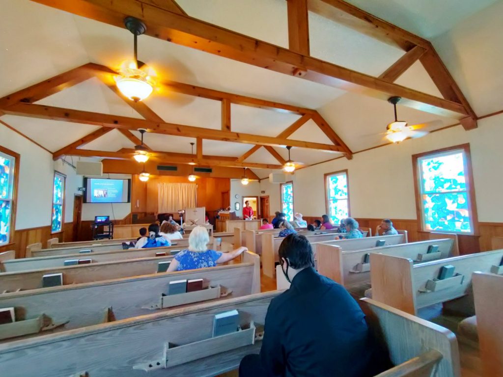 Shot from the back of the sanctuary as someone comes in through the door front right. Others are already seated. Front left sits a cart with laptop and projector aimed at a screen on the wall.