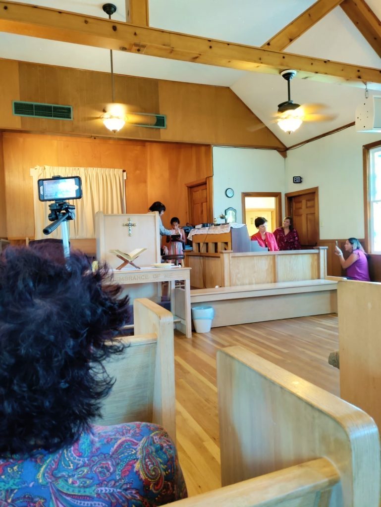 A shot from a few rows back in the sanctuary shows Priscilla helping Isabella get set up with microphone and iPad. Coser in, a woman is seen from behind aiming a phone on a tripod. Someone can be seen walking through the door at the front right of the room.