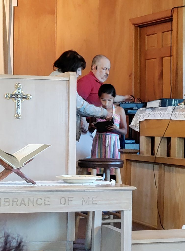 A little grirl in a dress sings while holding an iPad. Priscilla can be seen around the edge of the pulpit holding a clip-on mic to the girl's face. The pastor is behind her making adjustments on the sound board that is on a cart.