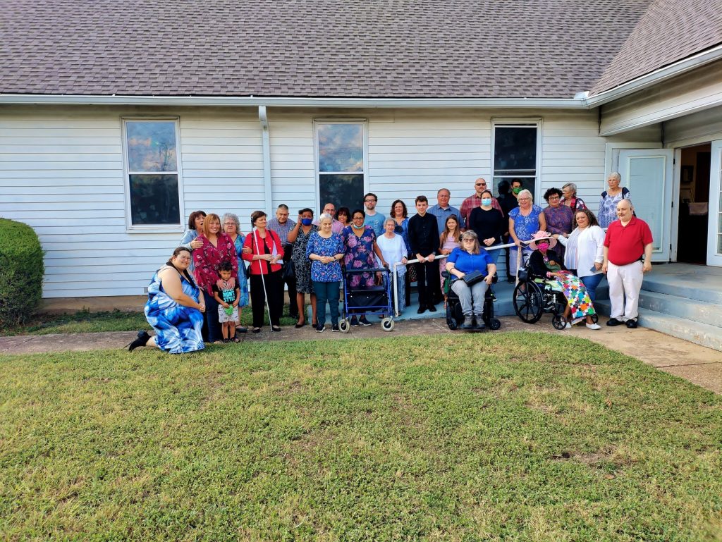 People are line up along the outside of the church building on the sidewalk that parallels the wheelchair ramp to the side door.