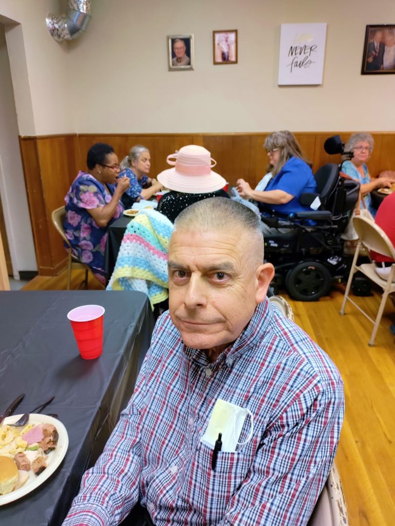 Don sits at a table, head lowered, a plate of food visible at the left of the frame. Behind him can be seen Pam in her chare and Ms. Fox's big white hat.