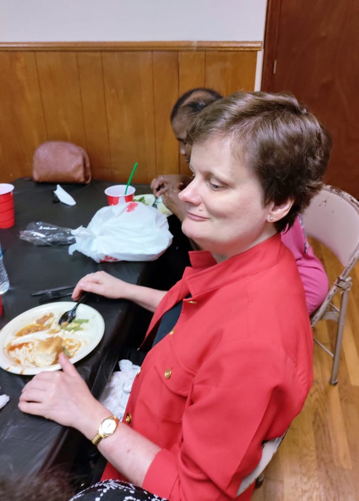 A woman with short, dark hair is pictured from the side with a plate of food on the table in front of her.