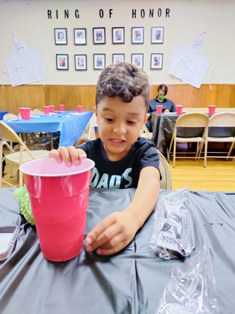 A small, DARK-HAIRED boy sits in the dining area holding a red plastic cup. He wears a dark t-shirt with partially obscured lettering that appeears to say "DADS" . Above him on a wall are several portraits with the words "RING OF HONOR" above them.