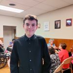 A teenaged boy poses in the middle of the room full of people seated at tables sharing a meal