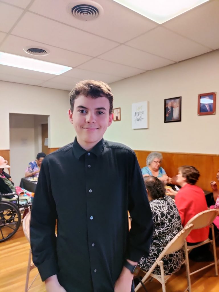 A teenaged boy poses in the middle of the room full of people seated at tables sharing a meal