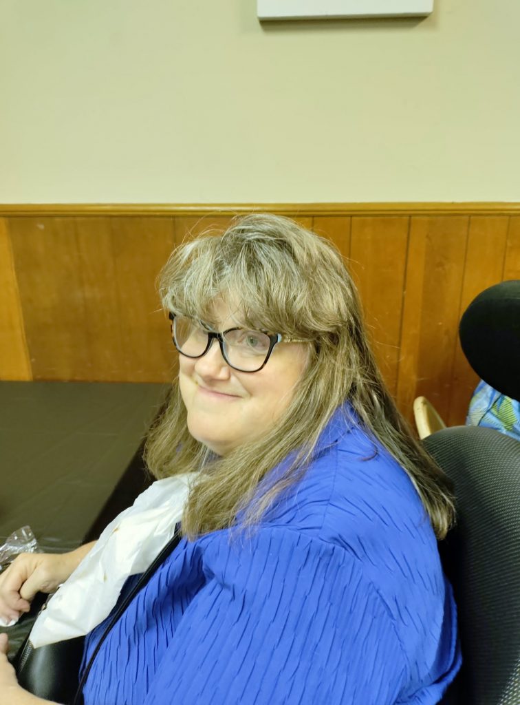 Pam, woman with long dark blond hair, wearing glasses and a blue top, seated in a chair