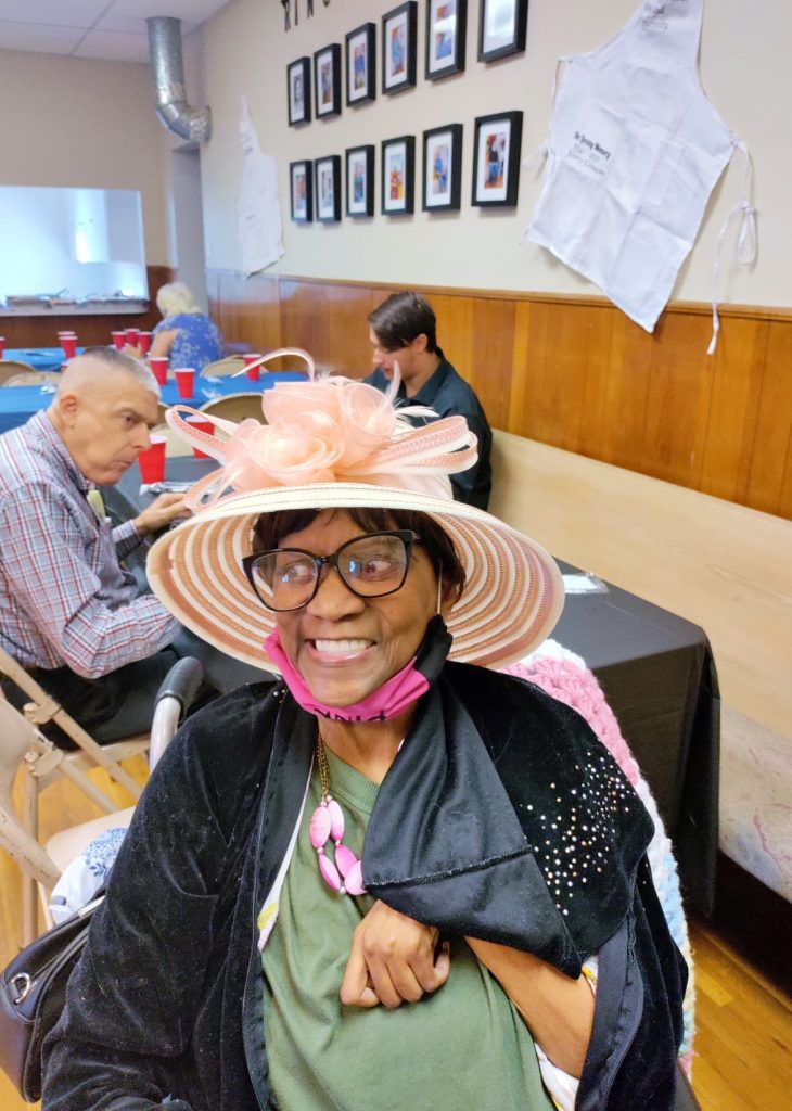 Ms. Fox, a dark skinned woman dressed in colorful clothing and wearing a big whate hat with bows